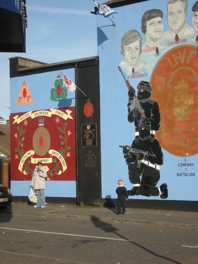 A woman holds her son's toy gun, behind is a UVF mural. Photo Vicky Richardson 