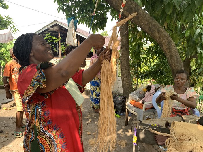 Female artisans in the frontyard of the Atelier in Masina, Kinshasa. Maman Monique is fixing a piece of raffia to be softened before weaving Jess Kilubu